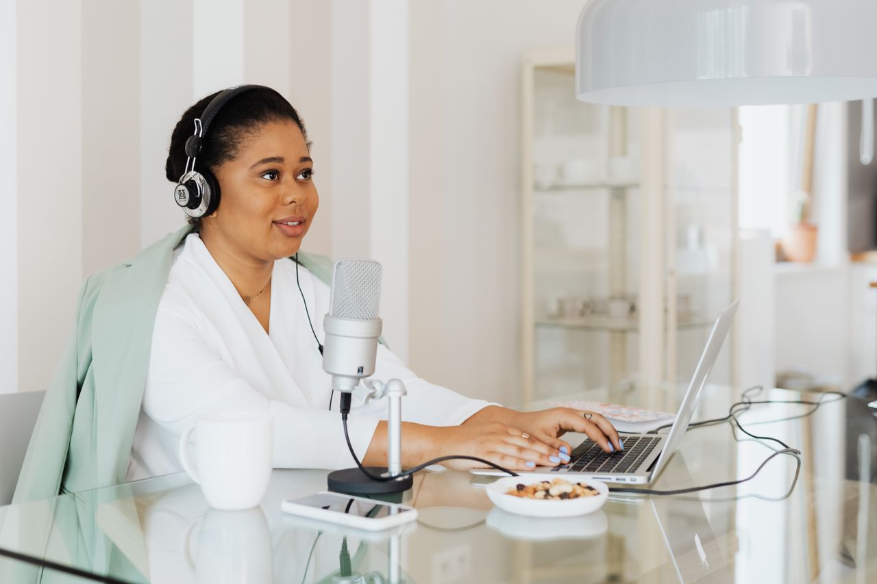 services-01 A woman working on a podcast in a modern home studio setup, using a laptop and microphone.